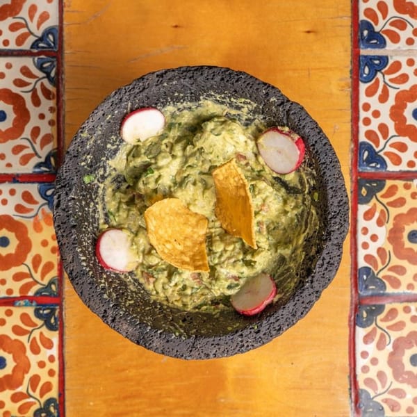 Don Pedro's guacamole in a molcajete with tortilla chips and radish slices on a colorful table.