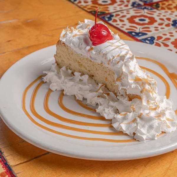 Slice of tres leches cake with whipped cream, caramel drizzle, and a cherry on a white plate.