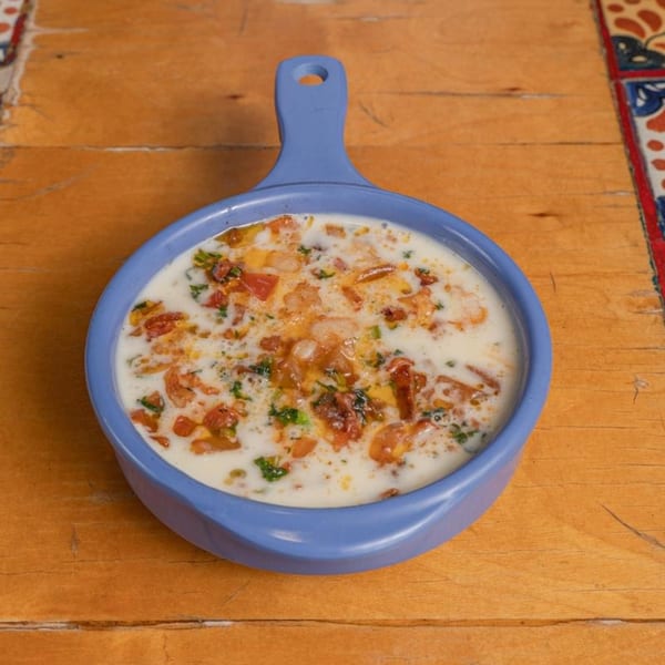A blue handled dish filled with creamy white queso, diced tomatoes, and green herbs on a wooden table.