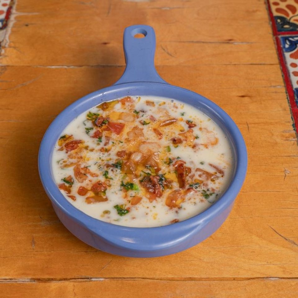 A blue handled dish filled with creamy white queso, diced tomatoes, and green herbs on a wooden table.
