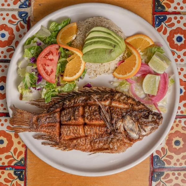 Whole fried fish with rice, avocado, salad, orange slices, and pickled onions on a white plate.